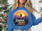 Wearing a Garment Graphics retirement sweatshirt, someone embraces unisex sizing with a blue shade featuring a hiker silhouetted against a sunset, mountains, and trees. The text reads Retired 2025, Not My Problem Anymore, set against the festive backdrop of a decorated Christmas tree.