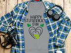 A Happy St. Patricks Day gray T-shirt by Garment Graphics, featuring a green shamrock design, rests on wood. Its surrounded by a blue and green plaid shirt, black camera, headphones, and a white coffee cup for a festive vibe.