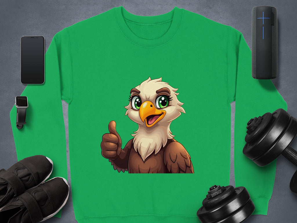 The Garment Graphics sweatshirt, featuring a green cartoon eagle and unisex sizing, sits cheerfully among a smartphone, smartwatch, sneakers, portable speaker, and two dumbbells on a gray surface.