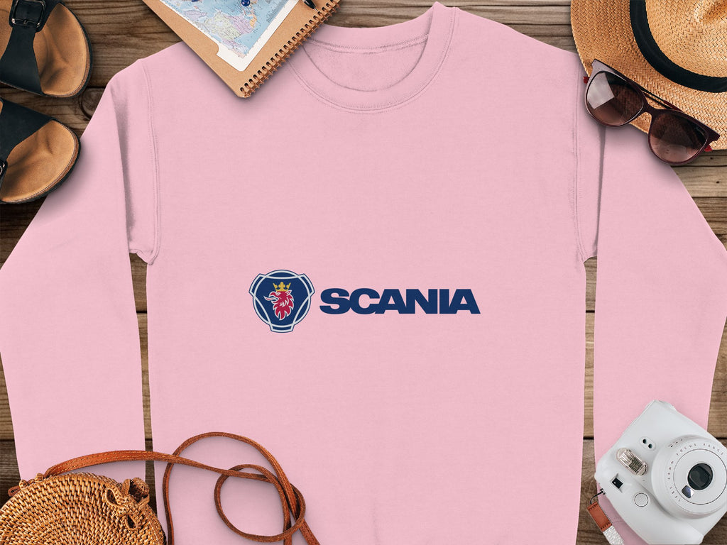 A pink Scania logo sweatshirt from Garment Graphics lies on a wooden surface, surrounded by a notebook, straw hat, sunglasses, camera, sandals, and a woven bag. Designed with unisex sizing in mind, its perfect for travel-ready casual outings and any adventure.