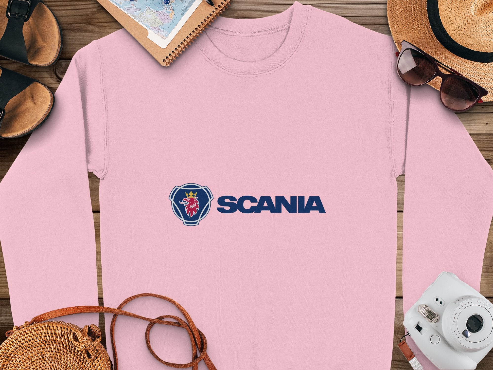 A pink Scania logo sweatshirt from Garment Graphics lies on a wooden surface, surrounded by a notebook, straw hat, sunglasses, camera, sandals, and a woven bag. Designed with unisex sizing in mind, its perfect for travel-ready casual outings and any adventure.