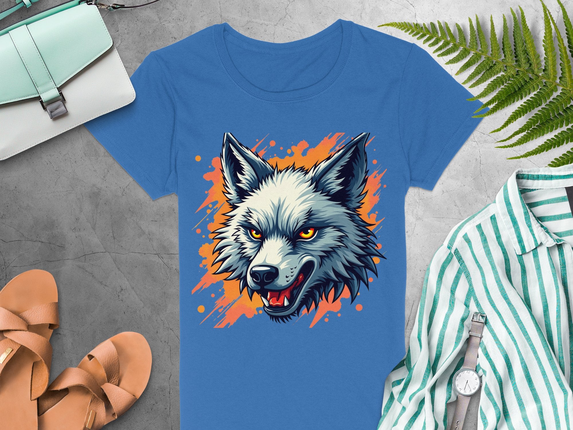 This 100% cotton blue T-shirt from Garment Graphics features a bold wolf print on an orange paint splash backdrop. Styled with a green plant, striped shirt, wristwatch, tan sandals, and light blue purse on a gray surface—perfect for showcasing your daring personality.