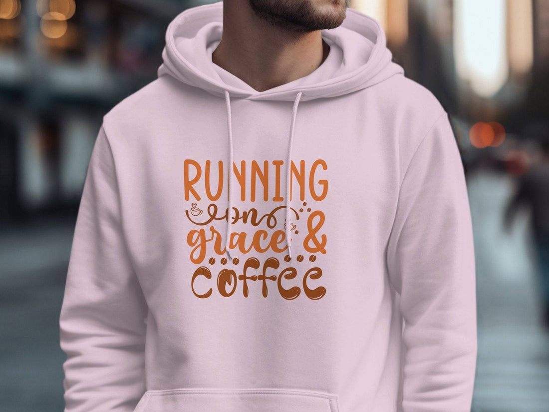 A classic fit black hoodie from Garment Graphics with Running on Grace & Coffee in orange lies on a wooden surface. Surrounded by Christmas decorations like pine cones, baubles, stars, and a steaming cup of coffee, this medium-heavy fabric piece exudes festive coziness.