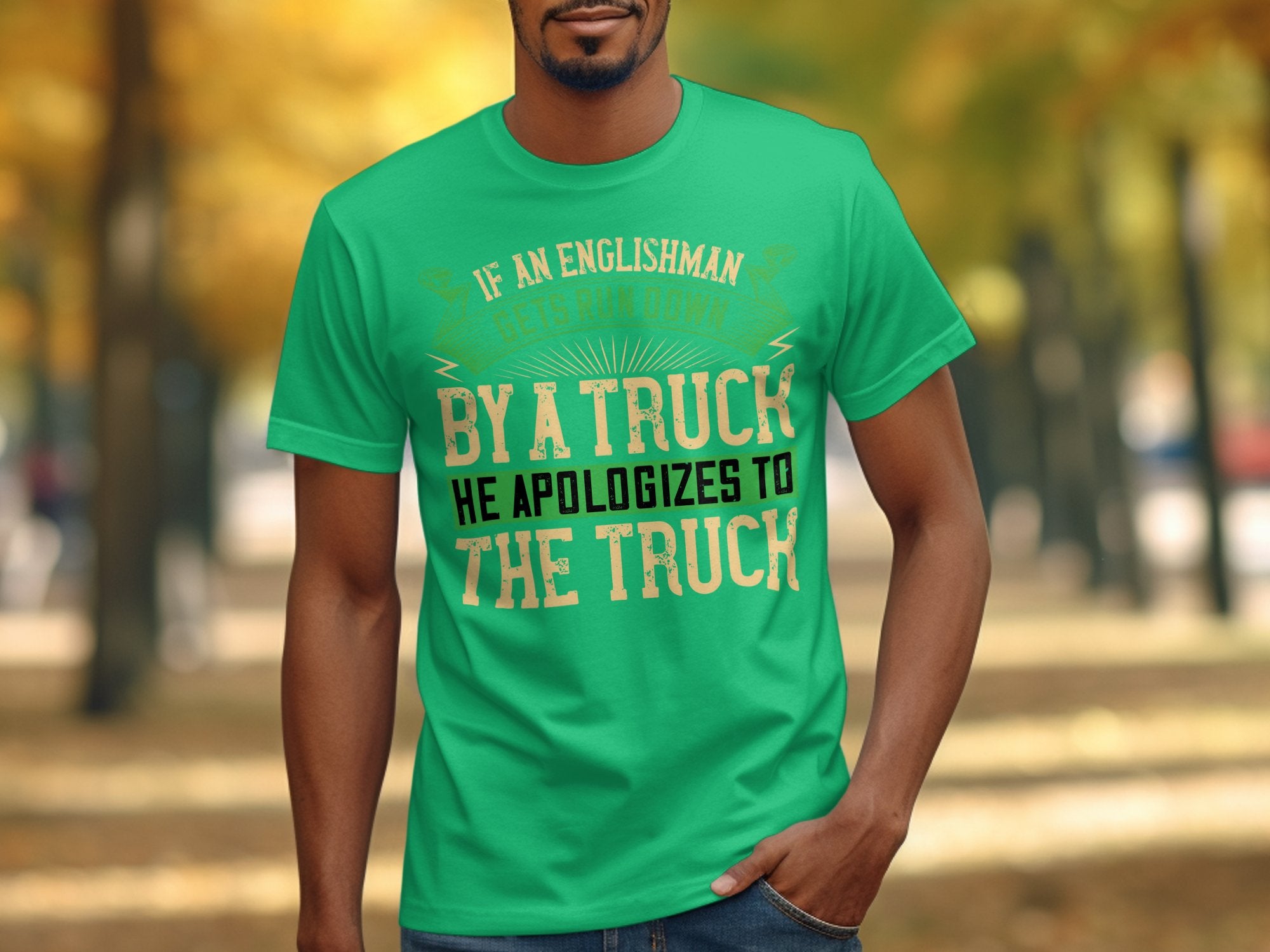 A person wearing a Garment Graphics green t-shirt with a witty design saying, If an Englishman gets run down by a truck, he apologizes to the truck, stands against a blurred park backdrop with trees and autumn leaves.