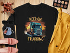 Garment Graphics offers a black graphic sweatshirt for truck enthusiasts featuring a bold truck design and the phrase KEEP ON TRUCKING. It is styled with pink shoes, a yellow jacket, sunglasses, makeup brushes, and an eyeshadow palette on a wooden surface.