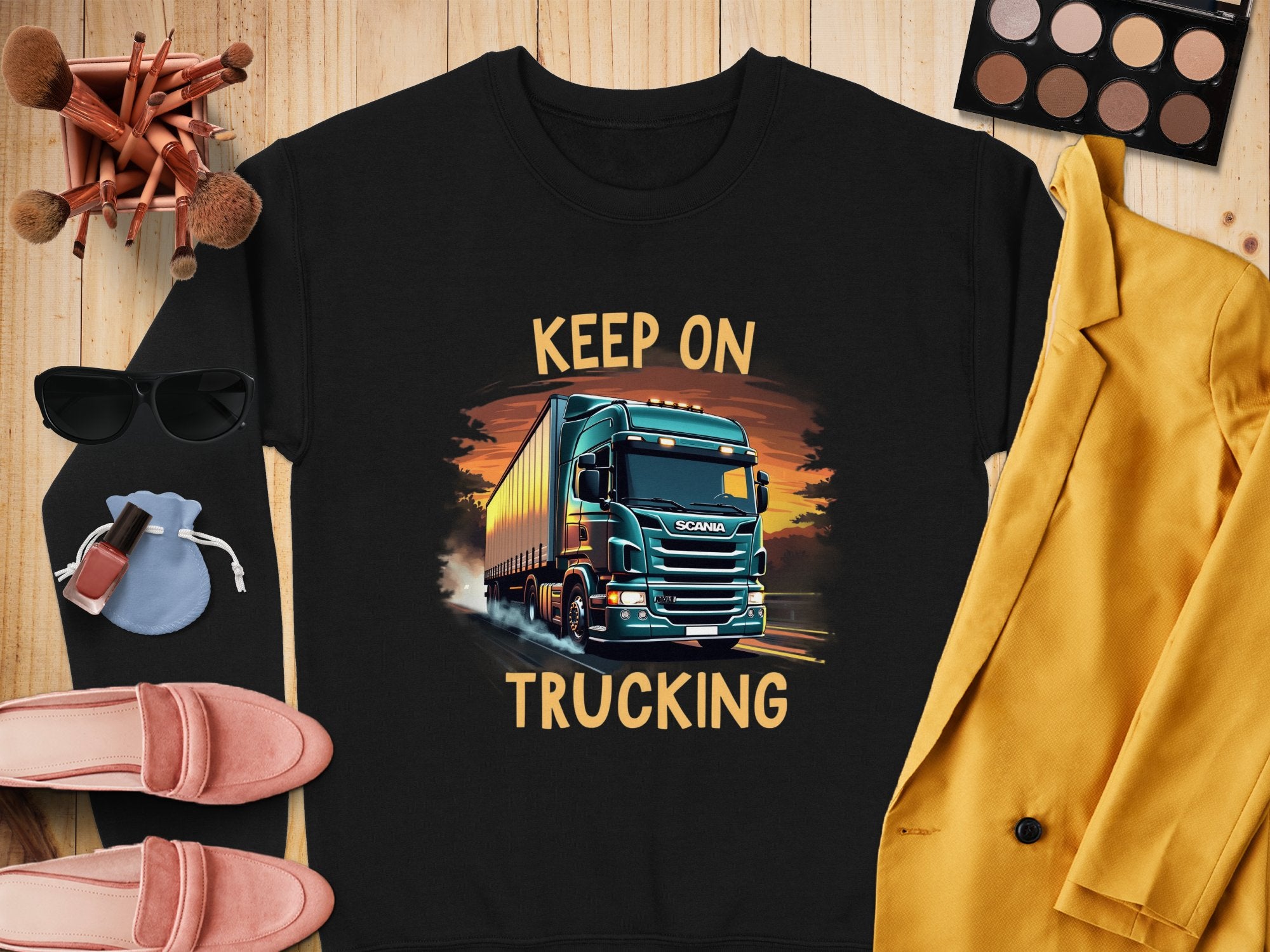 Garment Graphics offers a black graphic sweatshirt for truck enthusiasts featuring a bold truck design and the phrase KEEP ON TRUCKING. It is styled with pink shoes, a yellow jacket, sunglasses, makeup brushes, and an eyeshadow palette on a wooden surface.