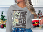 In a festive kitchen, a person holds a Santa-themed mug while wearing Garment Graphics’ gray sweatshirt featuring a cartoon alligator with a basketball and the phrase See You Later Alligator, surrounded by holiday decorations of ornaments and greenery.