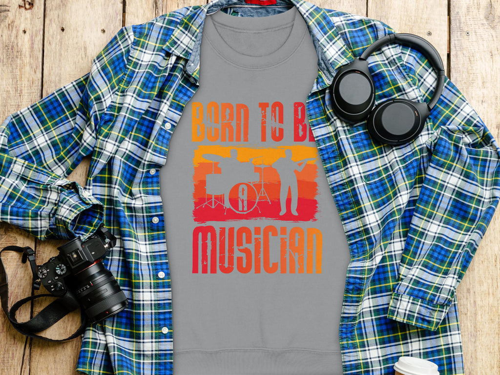Garment Graphics gray T-shirt, stating Born to be a Musician in bright orange and yellow, is perfect for music lovers. It features a microphone and instrument silhouettes against a blue plaid shirt with headphones, a camera, and a white cup on wood. Unisex sizing available.