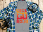 Garment Graphics gray T-shirt, stating Born to be a Musician in bright orange and yellow, is perfect for music lovers. It features a microphone and instrument silhouettes against a blue plaid shirt with headphones, a camera, and a white cup on wood. Unisex sizing available.