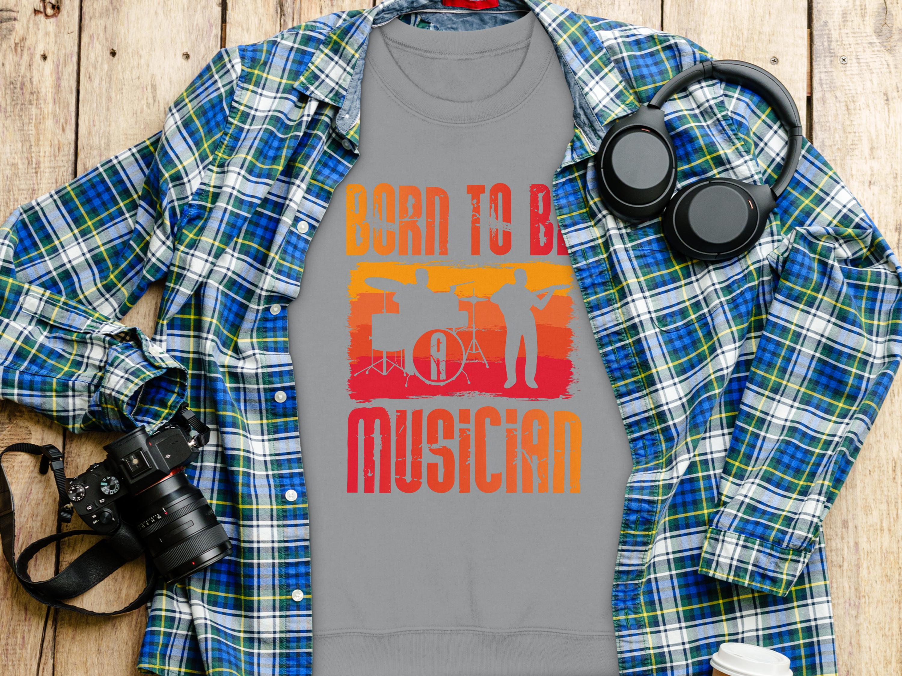 Garment Graphics gray T-shirt, stating Born to be a Musician in bright orange and yellow, is perfect for music lovers. It features a microphone and instrument silhouettes against a blue plaid shirt with headphones, a camera, and a white cup on wood. Unisex sizing available.