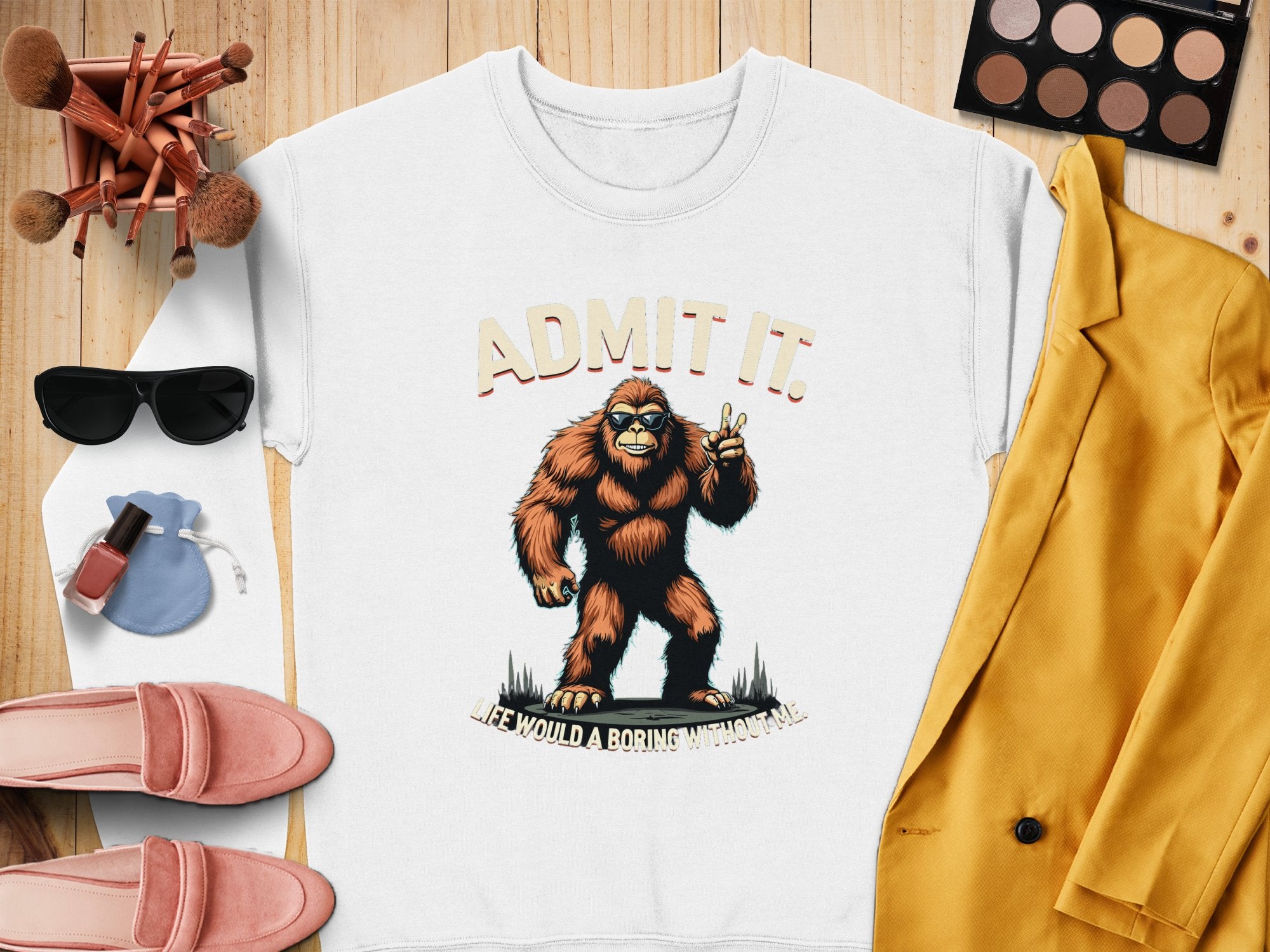 The Garment Graphics white sweatshirt showcases a Bigfoot character holding up two fingers with the slogan Admit it. Life would be boring without me. Its an ideal casual wear piece paired with a yellow blazer, pink loafers, sunglasses, makeup brushes, a pouch, and an eyeshadow palette.