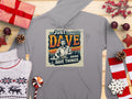 A Garment Graphics gray hoodie with medium-heavy fabric lays flat, showcasing a Just Dave Doing Dave Things graphic. Surrounded by wrapped gifts, pinecones, a sweater, red stars, and holiday decor on wood, it captures the festive spirit perfectly.