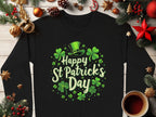 A Garment Graphics black shamrock graphic sweatshirt displaying Happy St Patricks Day with shamrocks and a green leprechaun hat sits on wood, surrounded by evergreen branches, pine cones, red stars, and a cup of tea, exuding festive cheer.