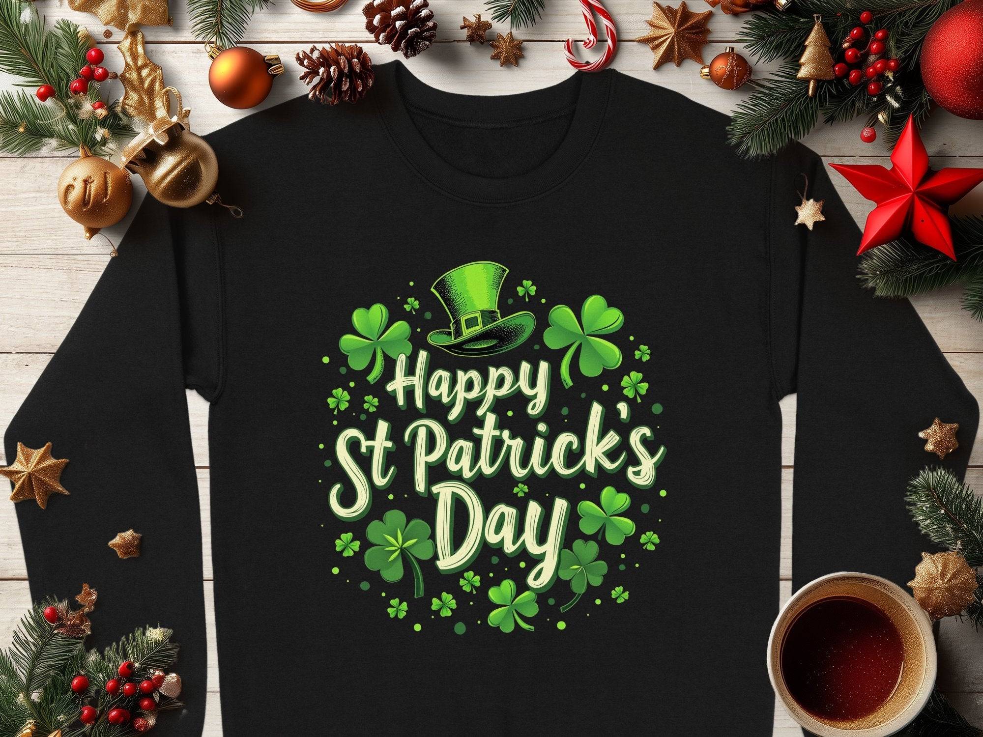 A Garment Graphics black shamrock graphic sweatshirt displaying Happy St Patricks Day with shamrocks and a green leprechaun hat sits on wood, surrounded by evergreen branches, pine cones, red stars, and a cup of tea, exuding festive cheer.