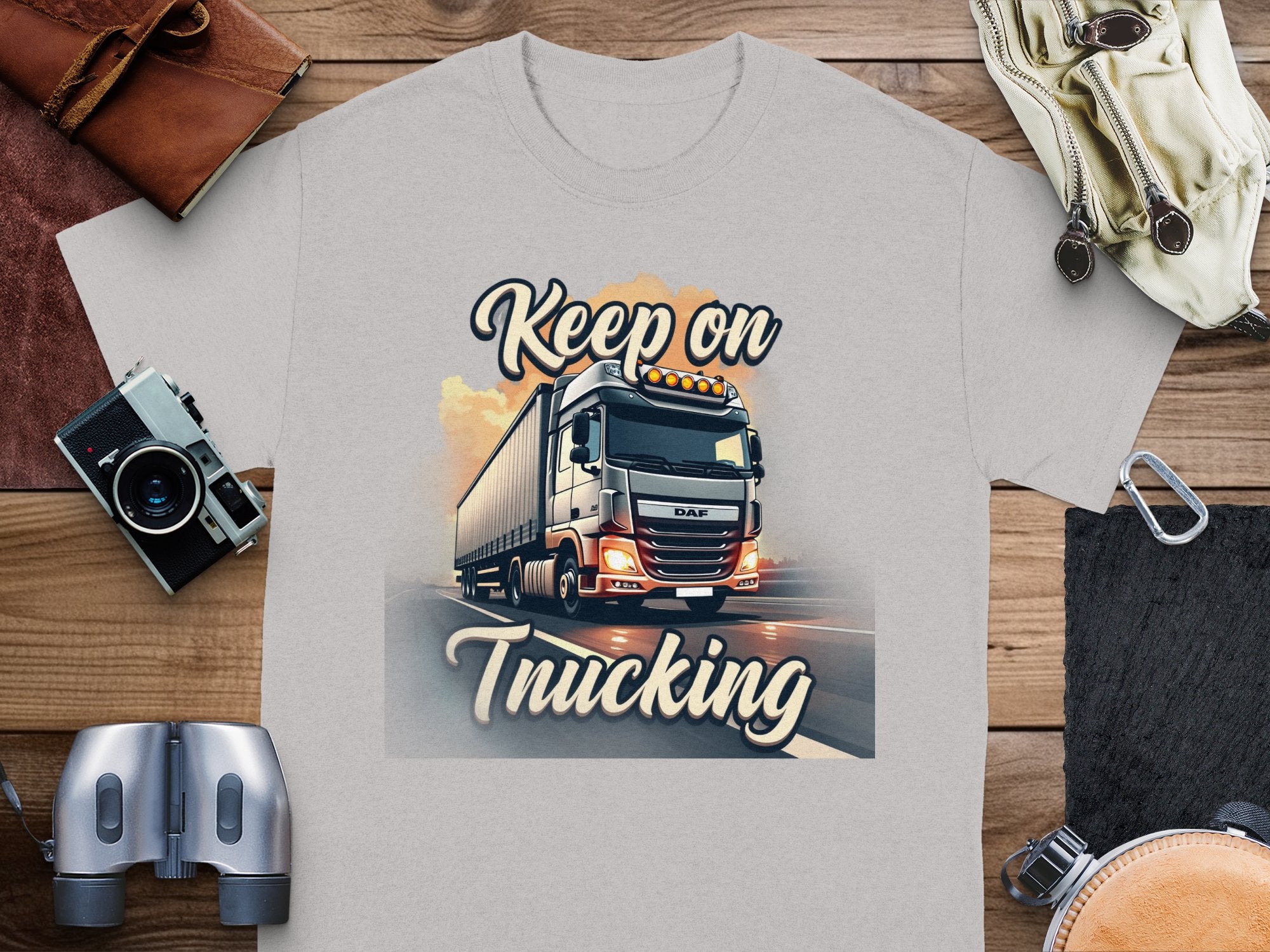 The Garment Graphics gray t-shirt features a dynamic large truck graphic on a highway, ideal for truck fans. Keep on Trucking is stylishly printed. Displayed on wood with a camera, binoculars, gloves, and wallet.