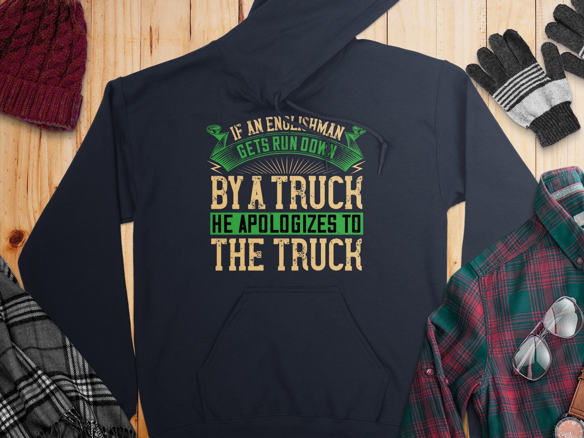 A Garment Graphics hoodie with the witty quote If an Englishman gets run over by a truck, he apologizes to the truck is displayed on a wooden surface, surrounded by a hat, gloves, flannel shirt, and other cozy clothing items.