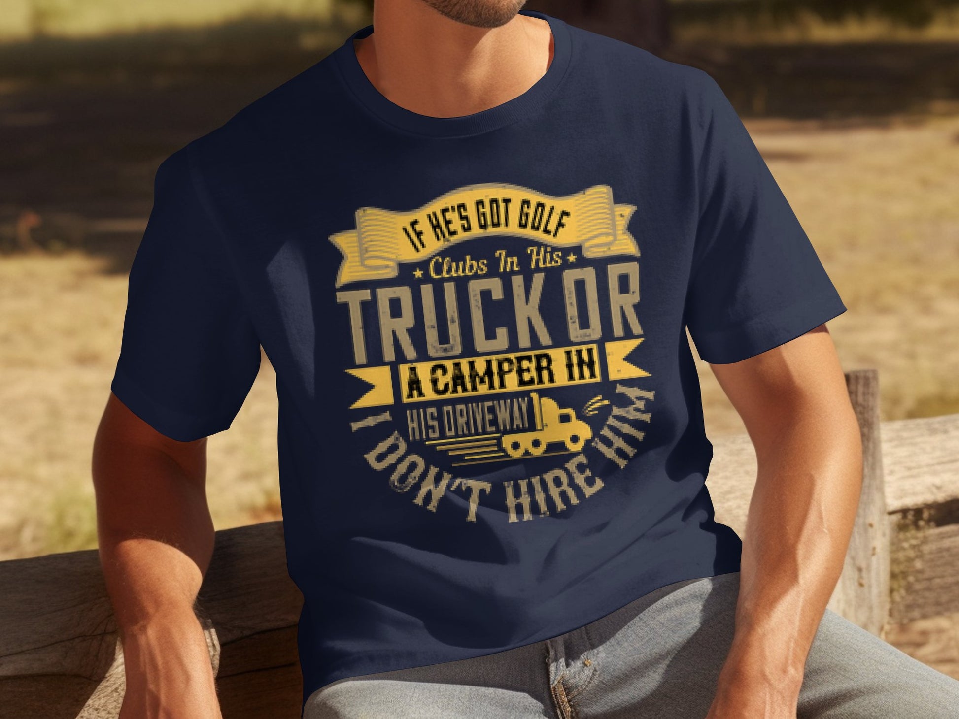 A person sits outdoors on a bench, wearing a navy blue t-shirt from Garment Graphics that humorously warns against hiring someone with golf clubs or camping gear, featuring playful graphics of trucks and campers.