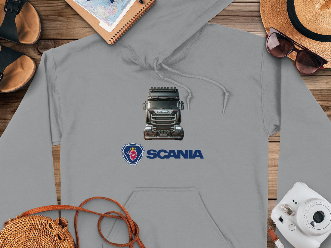 The Garment Graphics white hoodie features a graphic truck design with the Scania logo underneath. Ideal for truck enthusiasts, its displayed on a wooden surface alongside sandals, a notebook, sunglasses-topped straw hat, map, and camera.