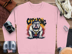 The Garment Graphics pink sweatshirt showcases an illustrated bulldog in a black cap, gold chain, and sunglasses for an urban vibe. City King appears above in graffiti style. Its laid on a wooden surface surrounded by adventurer gear like a camera and binoculars.