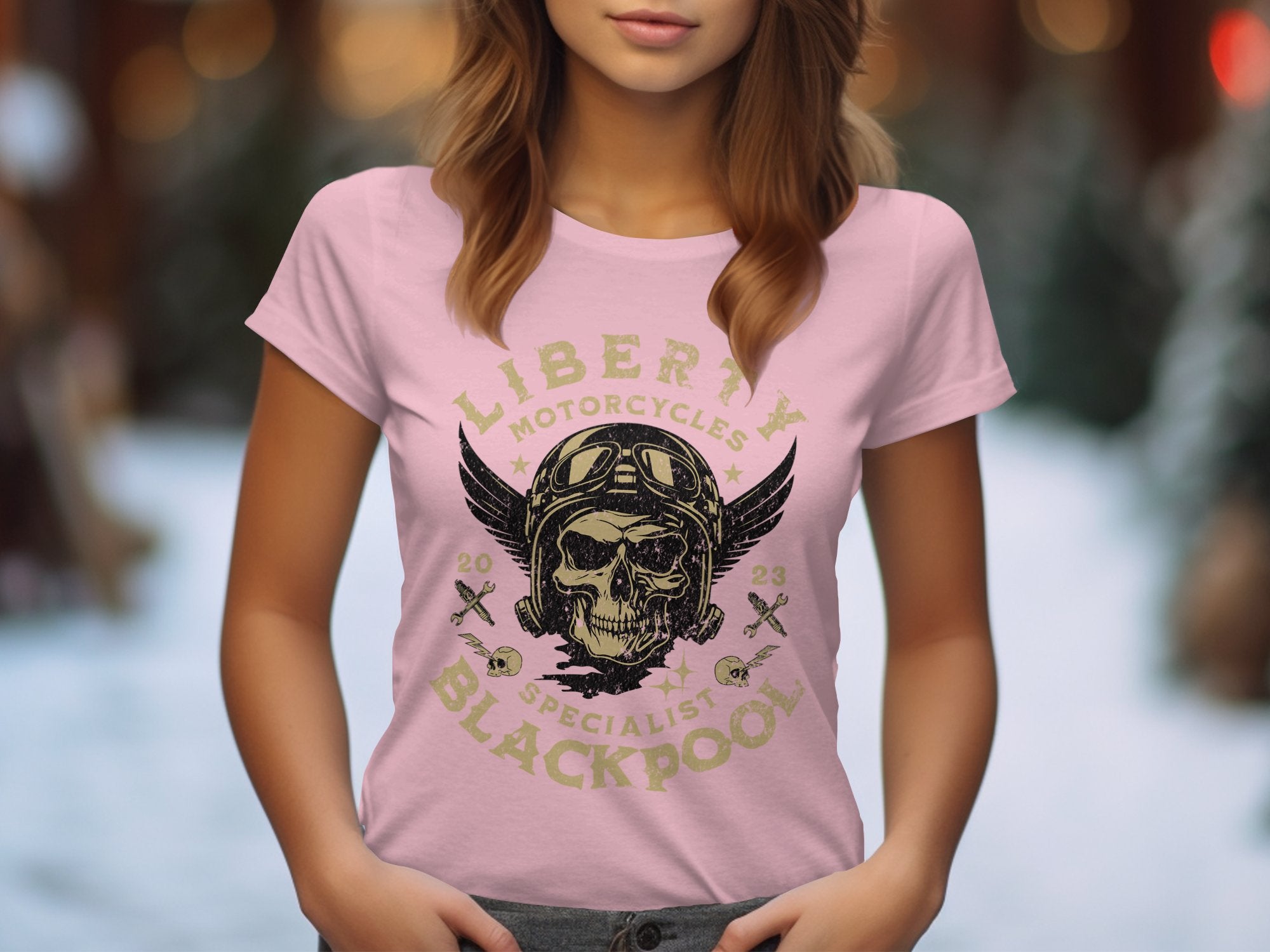 A woman poses outdoors in a pink Garment Graphics t-shirt featuring the renowned skull and wings design. The shirt reads Liberty Motorcycles Specialist Blackpool 2023, with a serene, tree-blurred background enhancing the tranquil vibe.