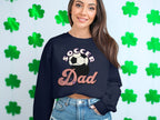 A woman with long brown hair smiles in a retro design black Soccer Dad sweatshirt by Garment Graphics, standing before a backdrop adorned with green shamrocks.