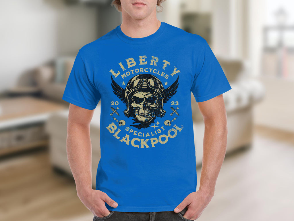 Sporting a vibrant blue T-shirt by Garment Graphics, the design is ideal for motorcycle fans, featuring a skull with wings, helmets, and crossed motorcycles. The text reads Liberty Motorcycles Specialist Blackpool 2023, set against a blurred indoor backdrop.