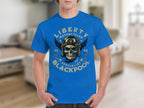 Sporting a vibrant blue T-shirt by Garment Graphics, the design is ideal for motorcycle fans, featuring a skull with wings, helmets, and crossed motorcycles. The text reads Liberty Motorcycles Specialist Blackpool 2023, set against a blurred indoor backdrop.