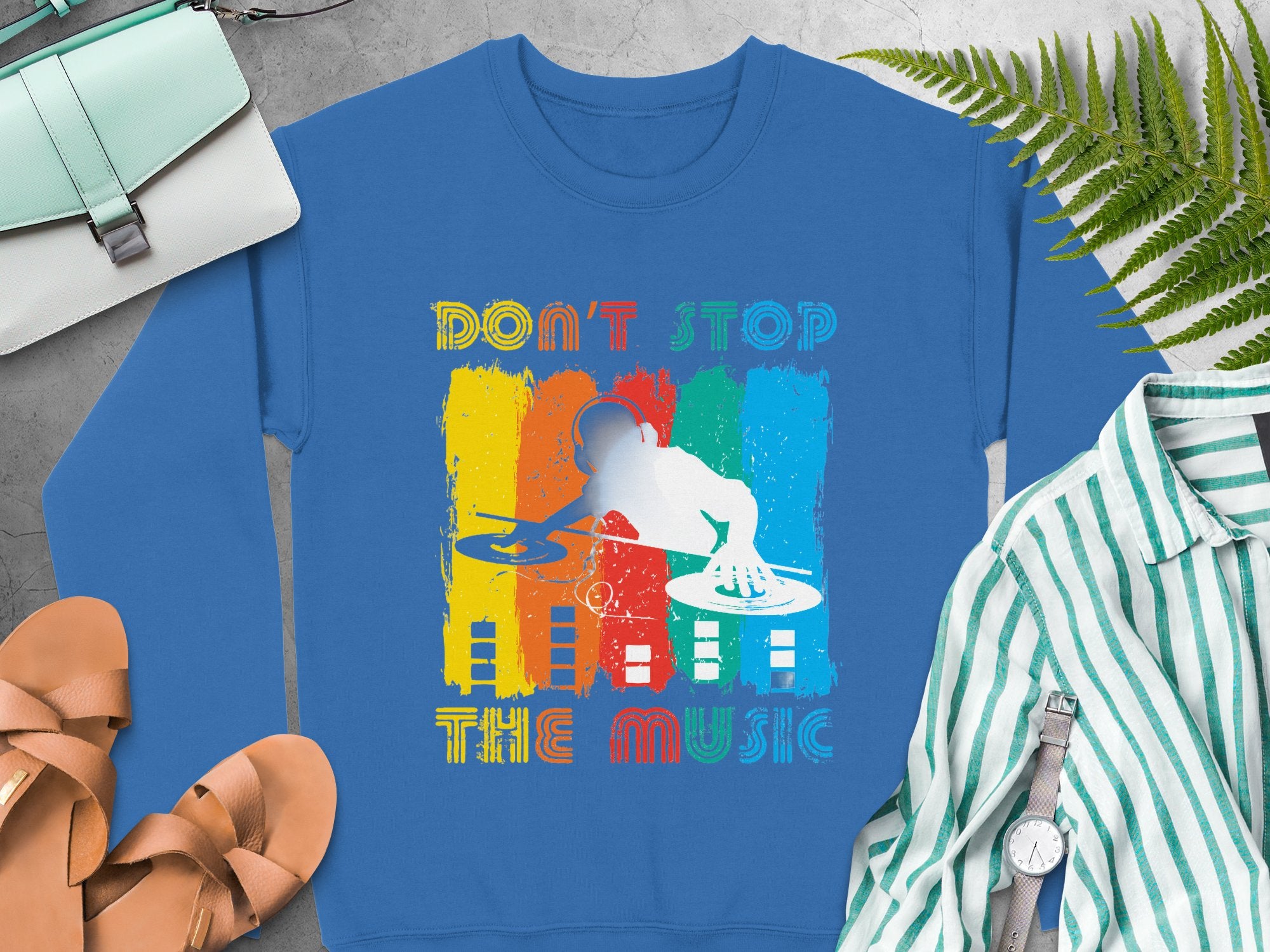Garment Graphics blue sweatshirt features a vibrant DJ design with Dont Stop the Music text, ideal for music lovers. It pairs beautifully with a striped shirt, sandals, fern leaves, and a light blue purse to complete the look.