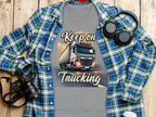 A Garment Graphics gray T-shirt with a bold Keep On Trucking graphic is layered over a blue plaid shirt. Nearby essentials include headphones, a camera, and a to-go coffee cup on wood, blending style and function in unisex sizing.