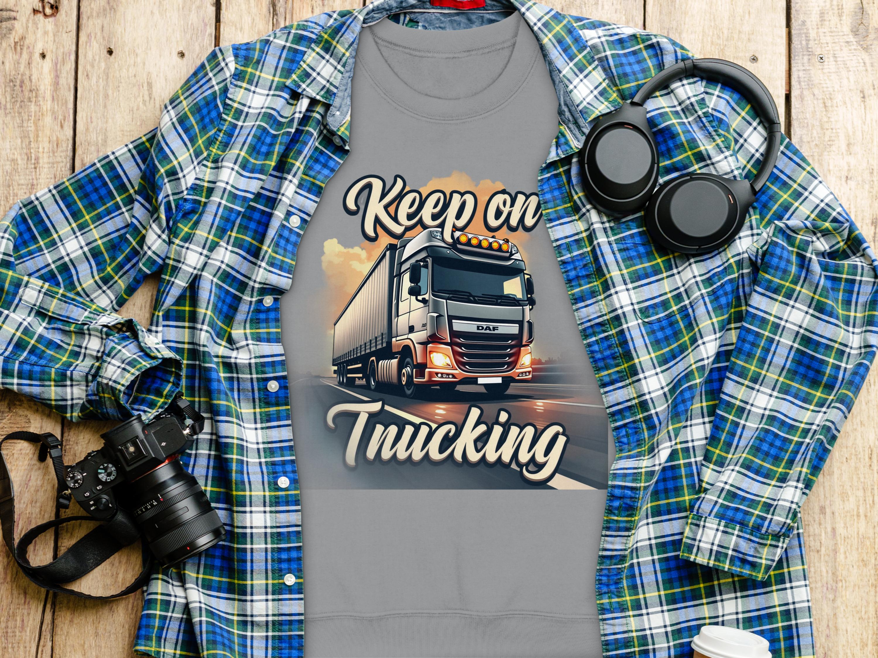 A Garment Graphics gray T-shirt with a bold Keep On Trucking graphic is layered over a blue plaid shirt. Nearby essentials include headphones, a camera, and a to-go coffee cup on wood, blending style and function in unisex sizing.