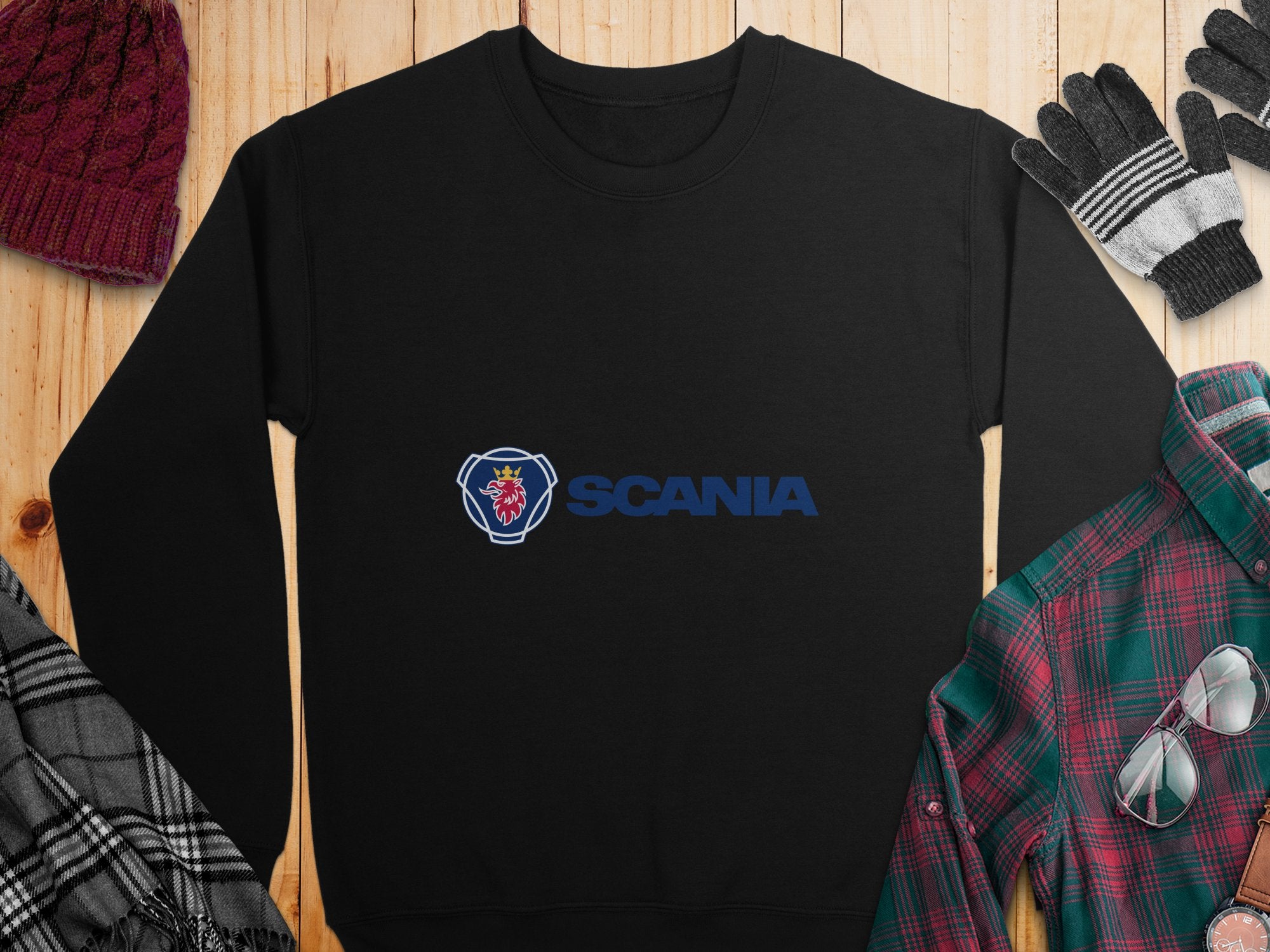 The Garment Graphics black sweatshirt with a griffin design and blue Scania logo is perfect for casual outings. It’s displayed on a wooden surface with a red knit cap, black and white gloves, plaid shirt, and scarf, offering unisex sizing for comfort.