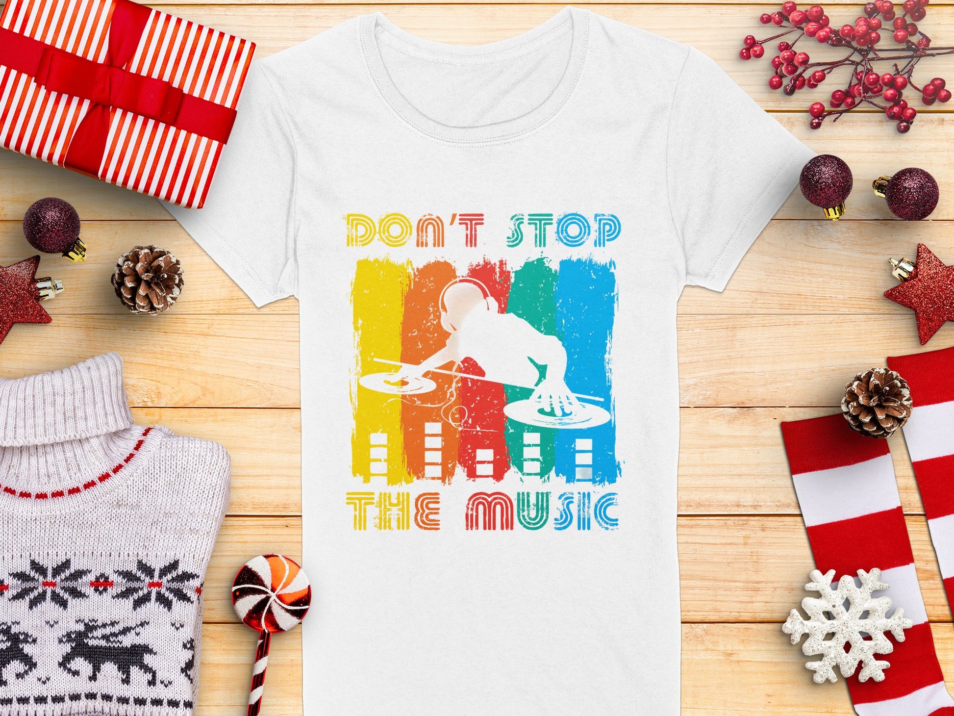 The Garment Graphics DJ graphic tee features Dont Stop the Music and a lively DJ design. It’s pictured with wrapped gifts, red berries, a candy cane, a Christmas sweater, and stars on wood—ideal for music lovers this festive season.