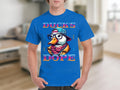 A person wears a blue Garment Graphics T-Shirt with a vibrant DUCKS DOPE design, showcasing a cartoon duck in a cap, glasses, and scarf. The background is a blurred room.