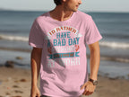 A person on the beach wears a light pink Garment Graphics fishing T-shirt with Id rather have a bad day fishing than a good day at work, perfect for anglers who love the sea.