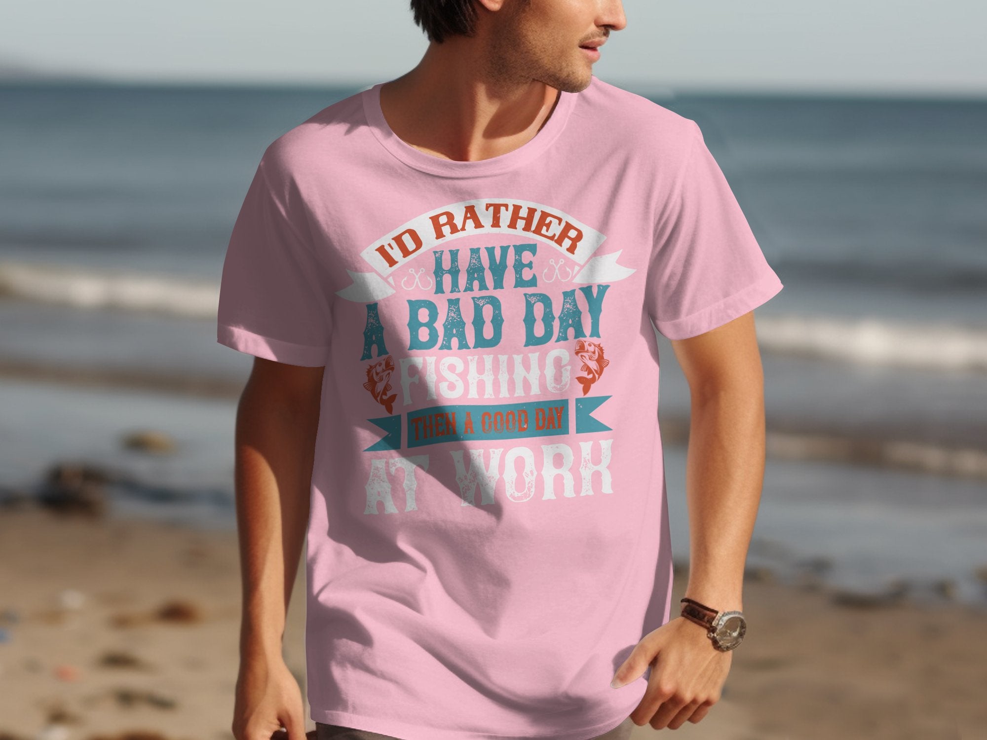 A person on the beach wears a light pink Garment Graphics fishing T-shirt with Id rather have a bad day fishing than a good day at work, perfect for anglers who love the sea.
