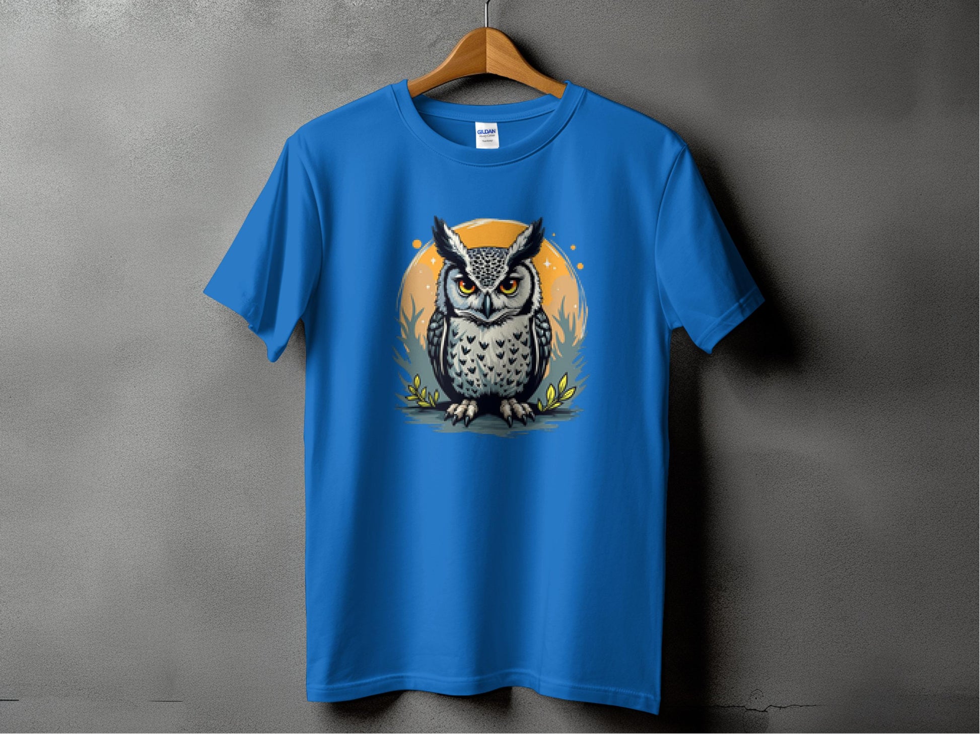 A Garment Graphics blue T-shirt showcases an owl with large, detailed eyes and feathers, set against an orange circle with plant motifs. The artistic design hangs gracefully on a wooden hanger.