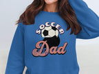 Wearing a Garment Graphics blue Soccer Dad graphic sweatshirt with a retro soccer ball and stars design, a person adjusts their hair, obscuring the lower half of their face.