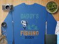 The Garment Graphics blue fishing sweatshirt features Daddys Fishing Buddy text with a delightful fish and person in boat illustration. Set on wood by a camera, lens, map, fruit bowl, and green book—ideal for fishing fans.