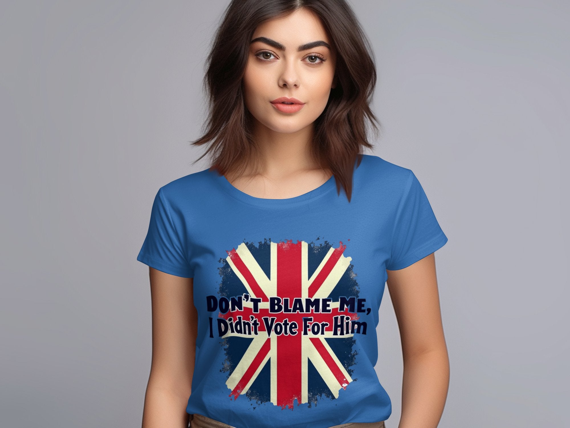 A woman with long brown hair wears a Garment Graphics blue T-shirt, 100% cotton, featuring the Union Jack and Dont blame me, I didnt vote for him text. She stands against a plain gray background.
