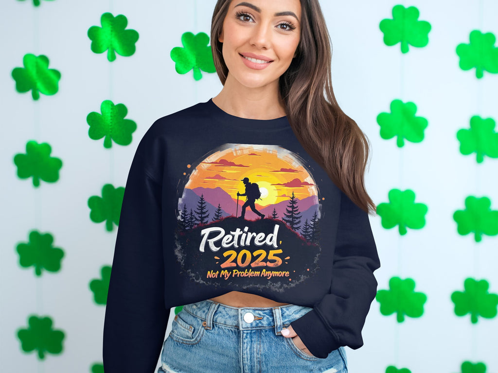 A smiling woman with long brown hair wears a black Retired 2025: Not My Problem Anymore unisex sweatshirt by Garment Graphics, featuring a hiker and sunset design. She stands before a wall decorated with green four-leaf clovers.