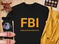 On a wooden surface is a black Garment Graphics sweatshirt with big yellow FBI and smaller Female Boob Inspector text, showcasing unisex sizing. Nearby are sunglasses, makeup brushes, a jacket, makeup palette, pink shoes, and a small blue pouch.