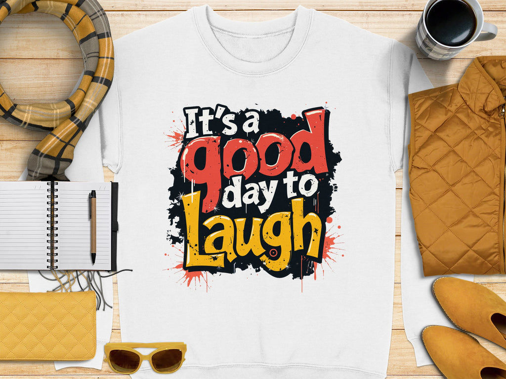 Flat lay featuring a white Garment Graphics unisex t-shirt with Its a good day to laugh in red, black, and yellow. The shirt is surrounded by a scarf, notebook, pen, sunglasses, mug, quilted jacket, and wallets on wood.