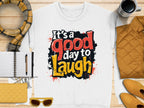 Flat lay featuring a white Garment Graphics unisex t-shirt with Its a good day to laugh in red, black, and yellow. The shirt is surrounded by a scarf, notebook, pen, sunglasses, mug, quilted jacket, and wallets on wood.