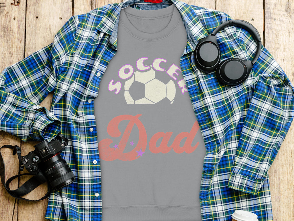 A flat lay features a Garment Graphics plaid shirt atop a gray Soccer Dad sweatshirt in unisex sizing. Nearby, headphones, a camera, and a partially visible coffee cup sit on the wooden surface.