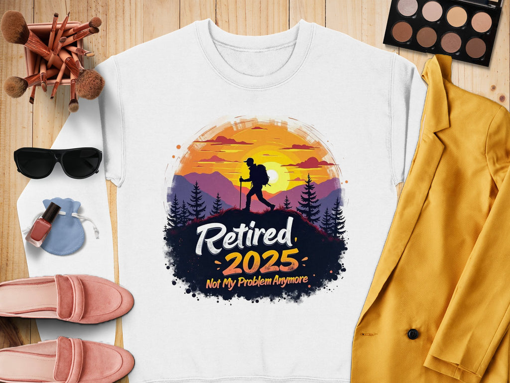 A flat lay features a Garment Graphics white retirement sweatshirt with a hiker design and Retired 2025. Not My Problem Anymore text, surrounded by pink shoes, a yellow blazer, makeup brushes, sunglasses, and an eyeshadow palette.