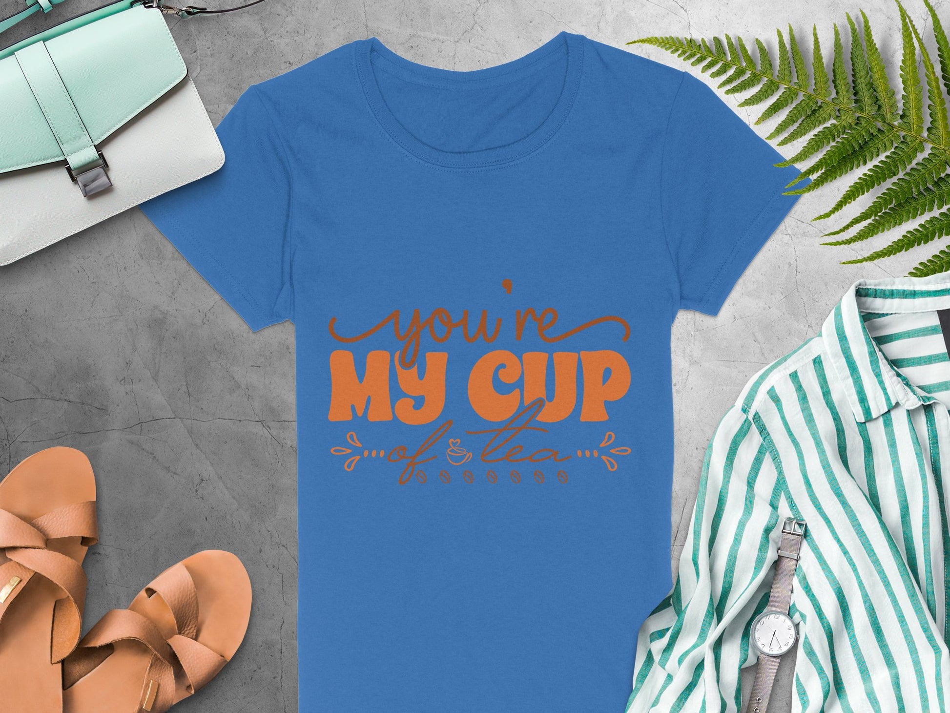 On a gray surface lies Garment Graphics blue 100% cotton T-shirt with Youre My Cup of Tea in orange lettering. Nearby are a green bag, tan sandals, a green and white striped shirt, a silver watch, and a fern leaf.