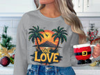 In a festive kitchen, a woman holding a Santa-shaped mug wears a grey Garment Graphics sweatshirt featuring the One Love Sunset design with palm trees and a vibrant sunset.