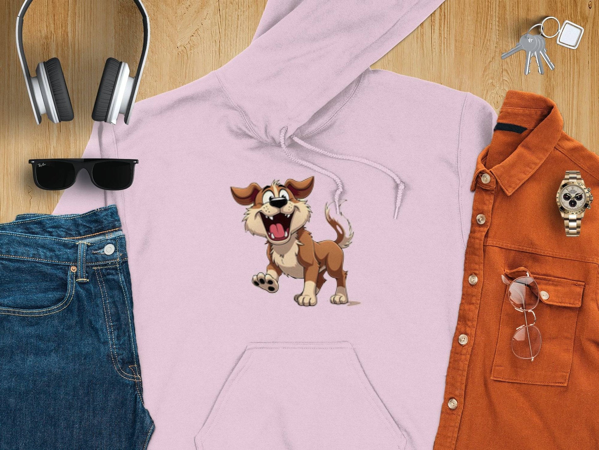 A Garment Graphics light pink cartoon puppy hoodie sits on a wooden surface, surrounded by headphones, sunglasses, blue jeans, an orange shirt, a wristwatch, and keys—ideal for stylish dog lovers.