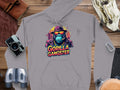 Garment Graphics presents the Gorilla Gangster gray hoodie, displaying a gorilla in a hat and sunglasses. Set against a wooden surface with a camera, binoculars, gloves, and notebook, this streetwear piece is ideal for those who love unique gangster designs.