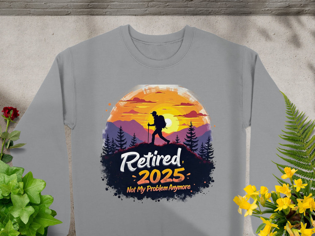Garment Graphics offers a gray unisex sweatshirt featuring a hiker silhouette against mountains and a sunset with Retired 2025, Not My Problem Anymore. The design, surrounded by plants and flowers on concrete, caters to outdoor lovers.